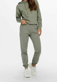 Olivgrön hoodie med dragsko, cropped design, kombinerad med matchande joggers. Mjuk tyg, elastiska muddar och ankle hems; bärs med vita sneakers.