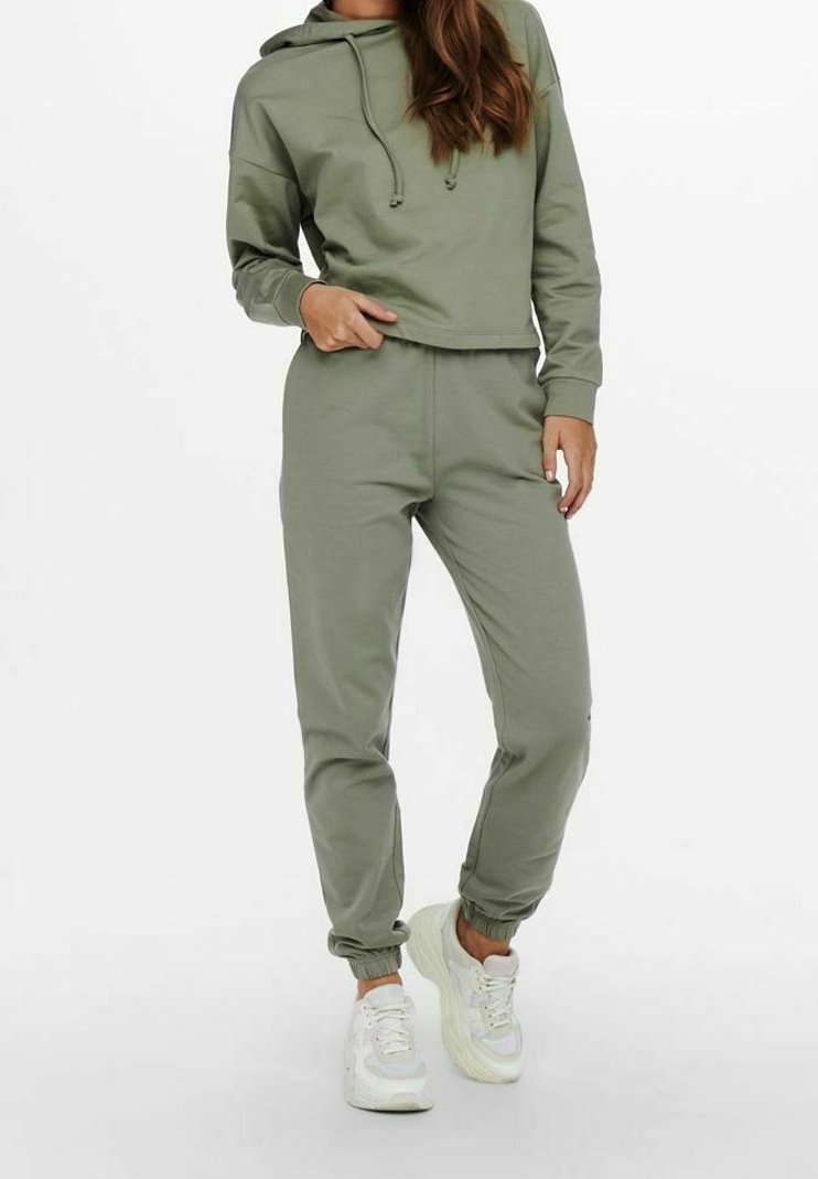 Olivgrön hoodie med dragsko, cropped design, kombinerad med matchande joggers. Mjuk tyg, elastiska muddar och ankle hems; bärs med vita sneakers.