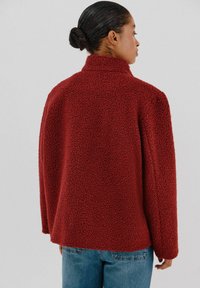 Rote strukturierte Jacke mit hohem Kragen, lockerer Passform und langen Ärmeln. Über blauen Jeans getragen, von hinten zu sehen.