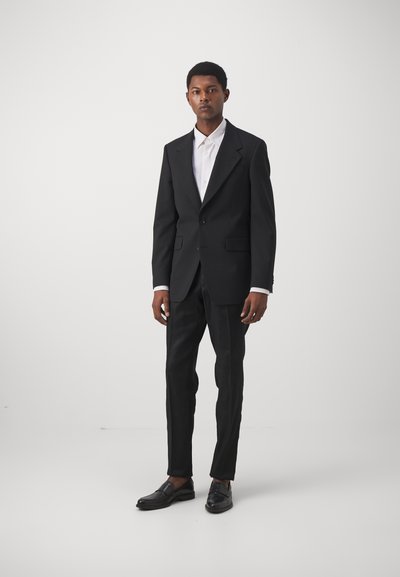 Zwart op maat gemaakt kostuum met een een-knops jasje en slim-fit broeken, gecombineerd met een witte overhemd met kraag en zwarte instappers.