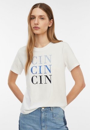 Młoda kobieta z długimi włosami, ubrana w białą koszulkę z nadrukiem "CIN" powtarzającym się trzykrotnie w różnych stylach oraz niebieskie dżinsy.