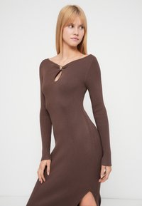 Brun ribbestrikket kjole med lange ermer, med en nøkkelhullsutringning og en dekorativ detalj. Designet inkluderer en sidesplitt for komfort.