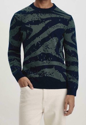 Mężczyzna ubrany w granatowy i zielony sweter z abstrakcyjnym wzorem z długimi rękawami oraz jasnobeżowe spodnie, jedna ręka w kieszeni.