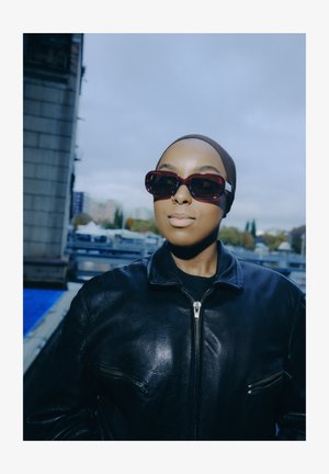 Črno usnjeno bomber jakno z žepki z zadrgo, kombinirano z bordo rdečimi sončnimi očali. Model nosi temno rutico, na ozadju z oblačnim nebom.