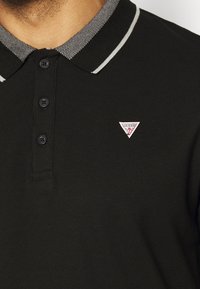 Czarny polo z teksturowanym kołnierzykiem z szarym wzorem, zapięciem na trzy guziki oraz małym, trójkątnym logo Guess na klatce piersiowej.