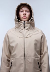 Beige vattentät jacka med hög krage och justerbar huva. Har en framzipper och sidofickor, slät, matt yta.