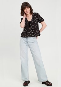 Blouse noire à imprimé floral, avec un col en V et un détail de nœud à l'avant, associée à un jean bleu clair à jambes larges et des sandales à plateforme noires.