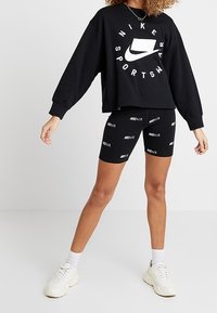 Black cropped sweatshirt with white Nike Sportswear logo, paired with black shorts featuring repeated "Nike Air" text. Neutral sneakers.