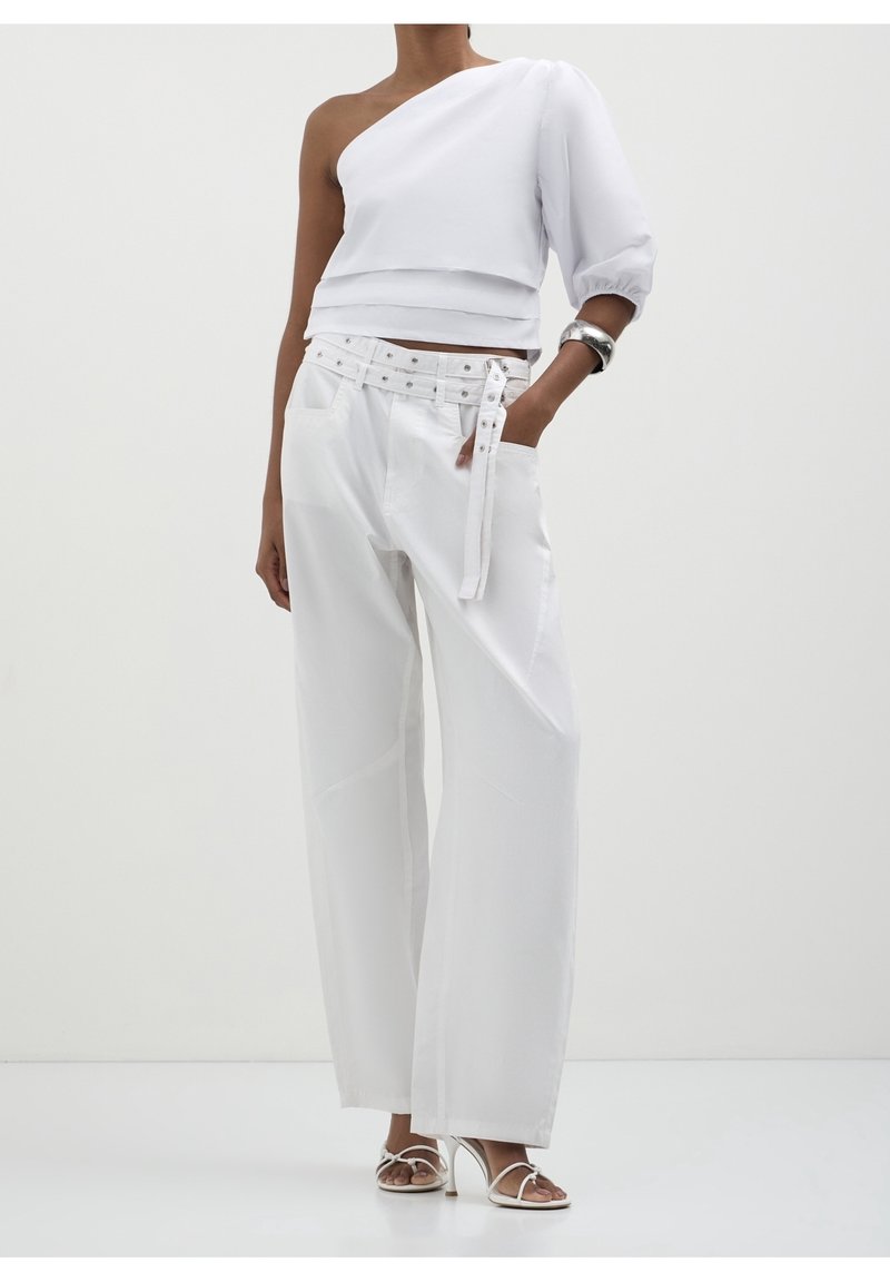 Femme portant un top blanc asymétrique, un pantalon à jambes larges avec ceinture cloutée, un bracelet argenté et des sandales blanches à talons hauts, debout devant un fond uni.