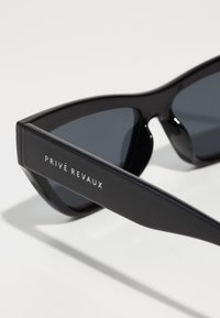 Ochelari de soare negri, în stil cat-eye, cu lentile întunecate, având un finisaj lucios și cu cuvintele "PRIVÉ REVAUX" gravate pe braț.