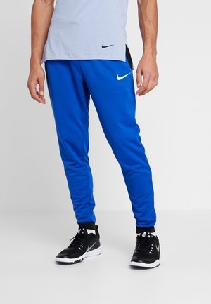 Homme portant un pantalon de jogging Nike bleu, des baskets Nike noires et blanches, et un t-shirt Nike bleu clair, debout sur un fond uni.