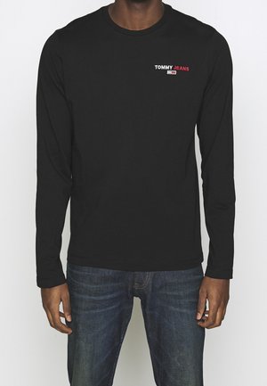 Zwart shirt met lange mouwen en een ronde hals, met een witte/rode "TOMMY JEANS"-logo op de borst, gecombineerd met donkerblauwe spijkerbroek.