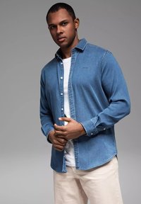 Helles blaue Denim-Hemd mit Knopfleiste und langen Ärmeln, kombiniert mit einem weißen T-Shirt darunter und cremefarbigen Hosen.