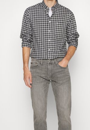 Homme portant une chemise à carreaux noir et blanc boutonnée avec les manches retroussées et un jean gris en denim, une main dans la poche, sur fond blanc.
