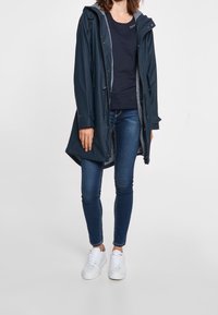 Vrouw draagt een marineblauw regenjas over een zwart shirt, blauwe skinny jeans en witte sneakers, staand tegen een effen lichte achtergrond.