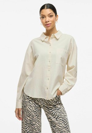 Light beige button-up shirt with a collar and a chest pocket, paired with patterned beige and gray zebra-print pants. Long sleeves rolled up.