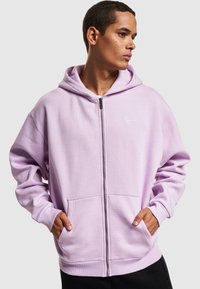 Lynlukkede hettegenser i lys lavendel bomullsblanding, med ensfarget design, frontlommer og ribbestrikket kant. Brodert logo på brystet.