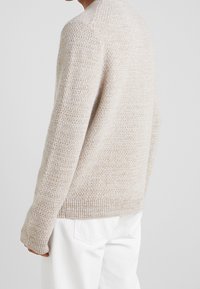 Beiger, gestrickter Pullover mit strukturiertem Muster, gerippten Bündchen und Saum. Kombiniert mit weißen Jeans, die einen lässigen Schnitt und entspanntes Design zeigen.