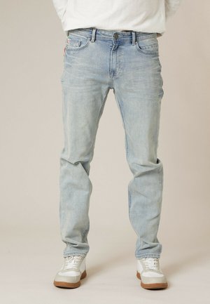 Homme portant un jean bleu clair et des baskets blanches, debout sur un fond neutre.