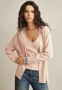 Cardigan rosa chiaro con una texture morbida, caratterizzato da una vestibilità rilassata e bottoni, indossato su un top a scollo a V coordinato, abbinato a jeans di denim.