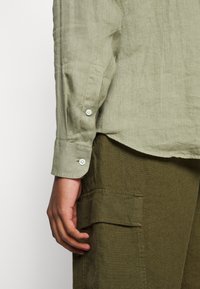 Bras gauche d'une personne portant une chemise vert clair à manches longues avec des boutons blancs et un pantalon vert foncé avec un rabat de poche latéral.