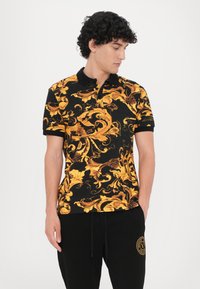 Camisa polo preta com padrão ornamentado em amarelo e castanho, mangas curtas e colarinho preto. Combinada com calças pretas com um logótipo.
