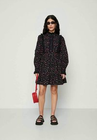 Vestito nero floreale con maniche a sbuffo lunghe e dettagli arricciati, abbinato a sandali neri e una piccola borsa rossa.