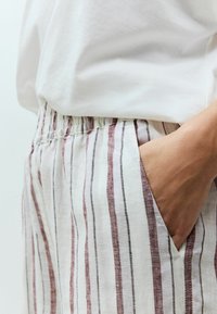 Pantaloni in lino a strisce con base bianca, caratterizzati da linee verticali rosse e nere. Elastico in vita e dettagli delle tasche laterali visibili.