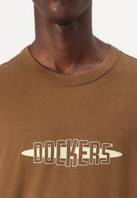 Brūns kokvilnas T-krekls ar lielu, bēšas krāsas "DOCKERS" logo uz krūtīm. Standarta apaļa kakla un īsas piedurknes. Gluda tekstūra.
