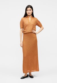 Nő áll, egyik kezét a csípőjén tartva, barna, rövid ujjú kötött felsőt és hosszú szoknyát visel, fekete balerinacipővel.