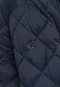 Marineblå quiltet dynejakke med diamantformede syninger, der har et rundt sort logoemblem og en glat, blank stoftekstur.