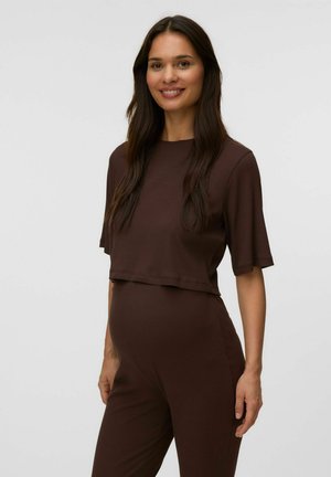 Lachende vrouw met lang donker haar, die een losse bruine top met korte mouwen en bijpassende hoge taille broek draagt tegen een effen lichte achtergrond.