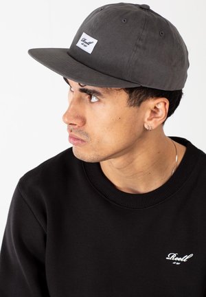 Black cap with a flat brim, featuring a white label on the front. Model wears a black sweatshirt with a small white logo on the chest.