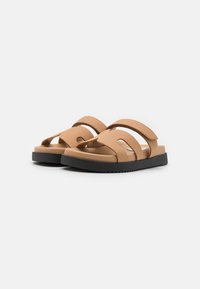Bruna slidesandal med två breda remmar, utskärningar och en texturerad svart gummisula. Designade för komfort och andningsförmåga.