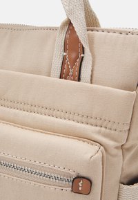 Bolsa de tela en beige claro con un acento de cuero marrón. Cuenta con un bolsillo con cremallera y un asa texturizada. Diseño compacto y funcional.