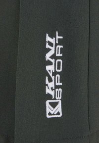 Schwarzer Stoff mit einem vertikalen, weißen gestickten Logo, das "KAN SPORTS" liest, sowie einem dazugehörigen Emblem auf der rechten Seite.
