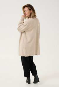 Béžový oversized svetr s dlouhými rukávy, měkkou strukturou a bočními štěrbiny, zkombinovaný s černými kalhotami a vysokými kotníkovými botami, na bílém pozadí.