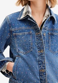 Jeansjacka med färgglada små nitar, bröstfickor och metallknappar, bärs över en rutig skjorta, hand i sidofickan, mot vit bakgrund.