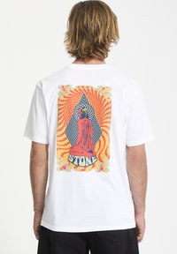 Hvid T-shirt med et farverigt grafisk design på ryggen, der viser en siddende figur med geometriske mønstre i rød, gul og sort.