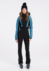 Zwart en teal skikostuum met een ceintuur in de taille, ritssluiting en lange mouwen. Accessoires zijn onder andere een zwarte gebreide beanie met een pompon.