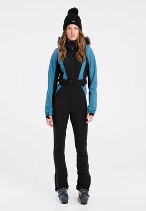 Zwart en teal skikostuum met een ceintuur in de taille, ritssluiting en lange mouwen. Accessoires zijn onder andere een zwarte gebreide beanie met een pompon.