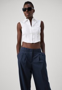Chemise blanche courte sans manches avec un plastron à l'avant, associée à un pantalon bleu marine à rayures avec plis et poches latérales. Lunettes de soleil noires.