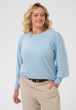 Femme blonde portant un chemisier bleu clair à manches longues et un pantalon beige, debout avec une main dans la poche et regardant sur le côté.