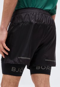 Άνδρας που φοράει μαύρα αθλητικά σορτς με στρώσεις και ελαστική μέση, με το όνομα της μάρκας να φαίνεται εν μέρει στο εσωτερικό στρώμα συμπίεσης.