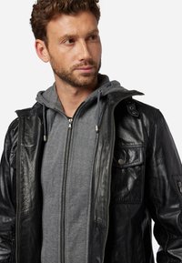 Schwarze Lederjacke mit silbernen Beschlägen, Reißverschluss vorne und geknöpften Taschen, über einem grauen Hoodie mit Reißverschluss und Kordelzügen getragen.