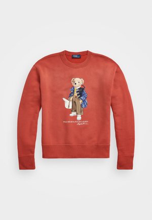 Bluza w kolorze rdzawym z nadrukiem przedstawiającym stojącego niedźwiedzia w niebieskiej kurtce, beżowych spodniach, białych tenisówkach oraz trzymającego białą torbę.