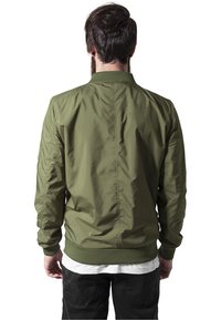 Olijfgroene bomberjack met een staande kraag, ribgebreide manchetten en onderrand, gladde stof en stikseldetails; gedragen met een wit shirt eronder.