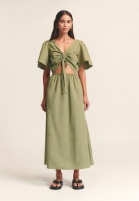 Olivově zelený dvoudílný outfit se skládá z krátkého upnutého topu s krátkými rukávy a maxi sukně s vysokým pasem, vyrobeného z lehkého materiálu.