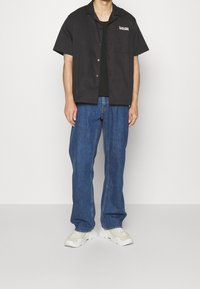 Maglietta nera a maniche corte con taschino e logo, abbinata a jeans blu a gamba larga e sneaker bianche chunky. Sfondo neutro.