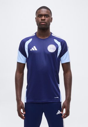 Muž stojaci čelom vpred oblečený v tmavomodrý a svetlomodrý športový dres a zodpovedajúce nohavice s logom Adidas na oboch.